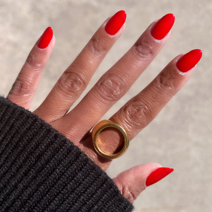 Hand with almond-shaped nails painted in vivid Lollipop quick dry polish wearing bold gold ring on neutral background