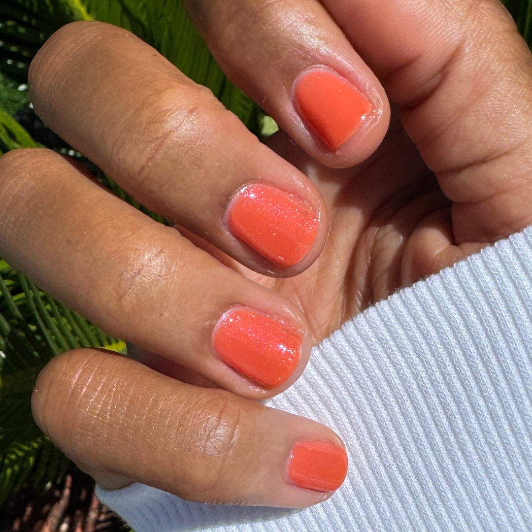 Hand with short nails painted in sheer red-orange I’ll Take Another Spritz gel polish with shimmer in natural light