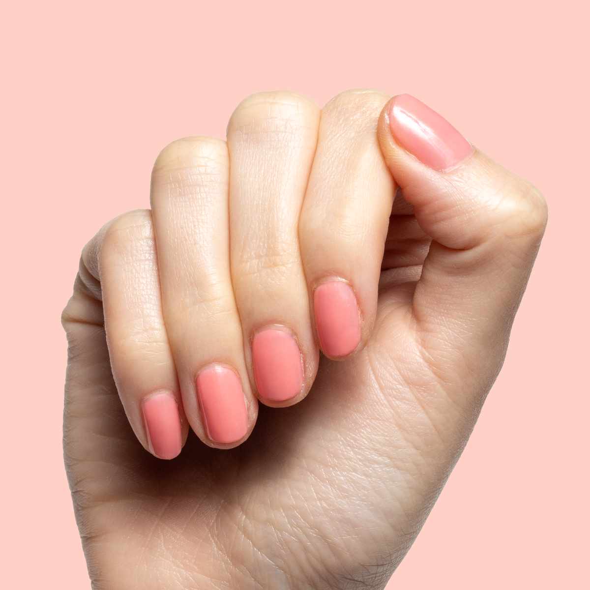 Hand with short natural nails painted in sheer coral pink using Papaya Gloss polish on soft pink background